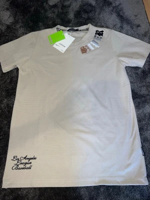 Beige t-shirt från Sevenstar - Snygg beige t-shirt från Sevenstar med diskret geometriskt mönster i tyget. Tryck i flera färger vid halsen och broderad text 'Los Angeles League Baseball' nertill. Perfekt för en trendig och avslappnad stil.