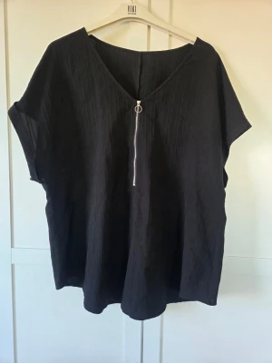 Svart v-ringad topp med dragkedja, XXL - Svart topp från med v-ringning och dekorativ dragkedja framtill. Modellen är loose fit med korta ärmar och lätt struktur i tyget. Perfekt för en avslappnad och stilren look.
