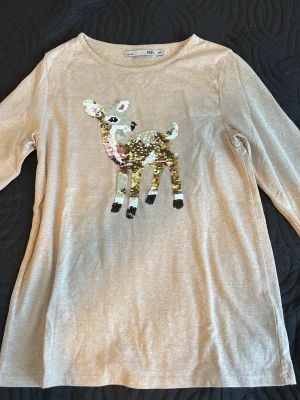 Beige långärmad topp med paljettmotiv - Beige långärmad topp med rund halsringning och ett stort paljettmotiv av ett rådjur framtill. Mjuk och skön bomullsblandning, perfekt för dig som gillar gulliga detaljer och lite extra glitter i vardagen.
