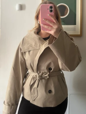 Beige trenchcoat från Gina Tricot - Stilren beige trenchcoat från Gina Tricot med dubbelknäppning, bred krage och knytskärp i midjan. Helt ny med prislapp, säljer pågrund av att den var lite stor på mig.