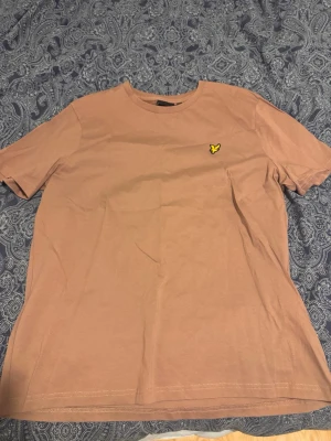 Beige t-shirt från Lyle & Scott - Säljer en beige t-shirt från Lyle & Scott med klassisk passform och korta ärmar. T-shirten har det ikoniska gula fågel-logotypen broderad på bröstet. Tillverkad i mjuk bomull, perfekt för en avslappnad stil.