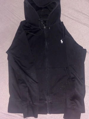 Svart hoodie från Polo Ralph Lauren - Svart hoodie med dragkedja från Polo Ralph Lauren. Klassisk design med vit broderad logga på bröstet, huva med dragsko och fickor framtill. Tillverkad i mjukt bomullsmaterial som är skönt att bära. Perfekt för en avslappnad och stilren look.