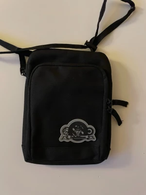 Sofialäktaren axelväska köpt på openair  - En svart axelväska i kompakt modell med justerbar rem och dragkedja. Väskan har ett tryckt märke framtill med motiv och flera fack för enkel organisering. Tillverkad i slitstarkt tygmaterial, perfekt för att bära det viktigaste.