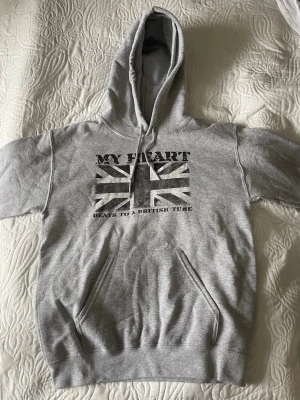 Grå hoodie från Drakenberg  med brittiskt tryck  - Säljer en grå hoodie med huva och känguruficka framtill. På bröstet finns ett tryck med brittiska flaggan och texten 'MY HEART BEATS TO A BRITISH TUNE'. Perfekt för dig som gillar streetstyle och vill ha en bekväm och stilren look.