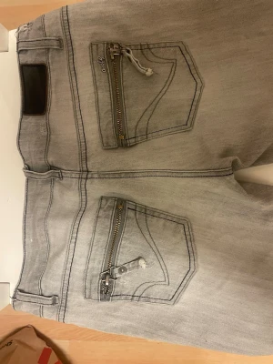 Grå slim fit jeans med dragkedjor - Snygga grå skinny  jeans med dragkedjor på bakfickorna och klassisk femficksdesign. Jeansen har en modern, smal passform och är tillverkade i denim med lätt stretch för extra komfort. Perfekta för en trendig och avslappnad look.