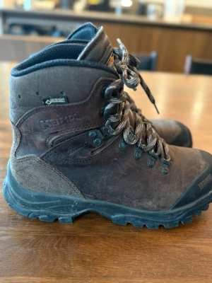 Bruna vandringskängor från Meindl - Stabila och slitstarka bruna vandringskängor från Meindl med Gore-Tex-membran. Kängorna har högt skaft, kraftig sula och tåliga metallhakar för snörning. Tillverkade i läder med detaljer i svart. Perfekta för utomhusaktiviteter och vandring i alla väder