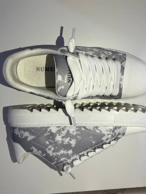 Vita och grå camo-sneakers från NUMERIS - Snygga sneakers från NUMER i vitt och grått camo-mönster. Skorna har vita snören, tåparti och sula samt coola silverfärgade nitar längs sidan. Ovandelen är i textil och tån i läderimitation. Perfekta för dig som vill sticka ut med din stil.