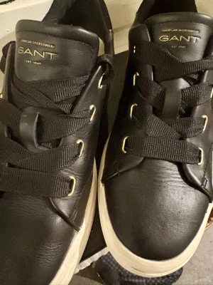 Svarta sneakers från GANT i läder - Stilrena svarta sneakers från GANT med ovandel i läder och breda svarta skosnören. Skorna har vit, chunky sula och guldfärgade detaljer vid snörningen samt GANT-logga på plösen. Perfekta för en clean och modern look.