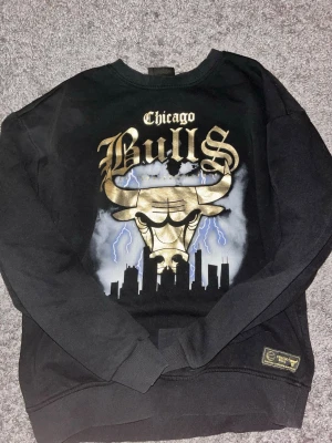 Chicago Bulls svart sweatshirt - Svart sweatshirt med stort Chicago Bulls-tryck i guld och vitt framtill. Motiv med tjurhuvud, blixtar och stadssilhuett. Rund halsringning och ribbade muddar. Perfekt för dig som gillar streetwear och basket. Den passar även bra om du har S