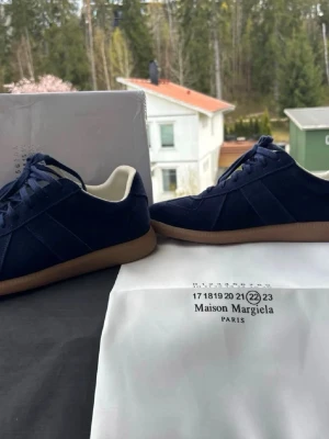 Maison Margiela blå sneakers i mocka - Snygga sneakers från Maison Margiela i mörkblå mocka med klassisk gummisula och vita detaljer på insidan. Skorna har lågt skaft och stilren design, perfekta för dig som gillar minimalistiskt mode och exklusiva material.