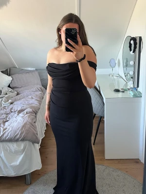 Svart offshoulder långklänning - Elegant svart långklänning med offshoulder-design och figurnära passform. Klänningen har en draperad överdel och är axelbandslös, vilket ger en stilren och modern look. Perfekt för bal eller festliga tillfällen. Köpt på Bubbleroom för 1199, aldrig använd och lappen hänger kvar 