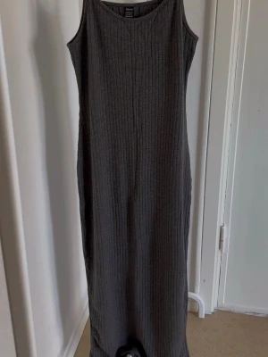 Grå ribbad långklänning  - En stilren grå långklänning i ribbad bomullsmix. Klänningen är ärmlös med rund halsringning och har en rak, kroppsnära passform. Perfekt för en enkel och avslappnad look.