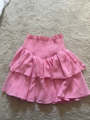 Rosa volangkjol med smock - Supersöt rosa kortkjol med bred smock i midjan och dubbla volanger. Kjolen har en luftig och flowy känsla, perfekt för varma dagar. Den stretchiga midjan gör den bekväm och enkel att bära. Passar perfekt till en enkel topp eller hoodie.
