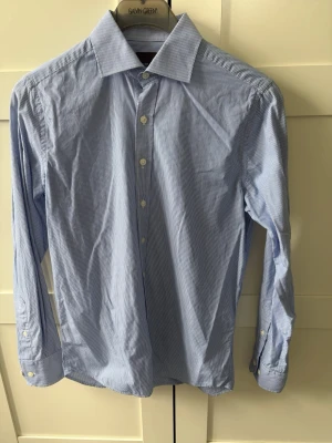 Blårandig skjorta från Oscar Jacobson - Säljer en klassisk blårandig skjorta från Oscar Jacobson i slim fit. Skjortan har långa ärmar, vit knappstängning och en snygg krage. Tillverkad i mjuk bomull som känns skön mot huden. Perfekt för dig som gillar stilrena och tidlösa plagg.