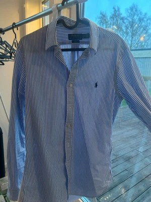 Blårandig skjorta från Polo Ralph Lauren - Inga deffekter på skjortan.                                        71 cm på längden.                                              Snygg blårandig skjorta från Polo Ralph Lauren i slim fit-modell. Skjortan har klassisk krage, långa ärmar och knäppning framtill. Ikoniska loggan broderad på bröstet. Perfekt för en stilren look.
