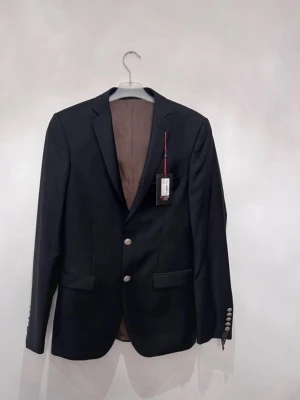 Svart kavaj  - Stilren svart kavaj med två silverfärgade knappar framtill och fem dekorativa knappar vid ärmsluten. Kavajen har klassisk passform, bruna innerfoder och två fickor framtill. Perfekt för en dressad look. Storlek L 146