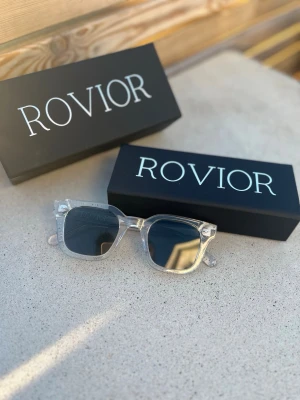 Klara solglasögon från Rovior - Snygga solglasögon från Rovior med 400 uv skydd transparent båge och mörka glas. Rektangulär form som ger en modern vibe och passar till många stilar. Kommer med originaletui och box. Perfekta för soliga dagar och en cool look. Nypris 699