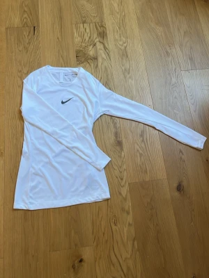 Vit långärmad träningströja Nike - Vit långärmad träningströja från Nike med svart swoosh-logga på bröstet. Tillverkad i ett lätt och stretchigt funktionsmaterial som andas, perfekt för träning eller sport. Tröjan har rund halsringning och en smal passform.