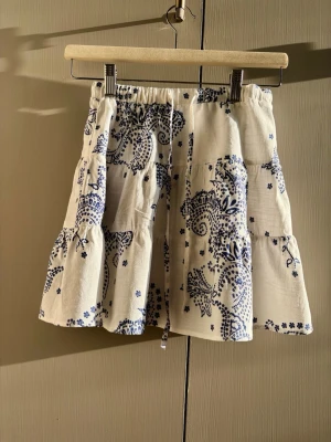 Vit och blå volangkjol med mönster - Supersöt kort kjol i vitt med blått blommigt mönster och volanger. Kjolen har resår i midjan och knytband framtill för extra passform. Perfekt för varma dagar och ger en avslappnad men trendig look.