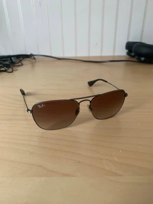 Ray-Ban bruna solglasögon aviator - Snygga Ray-Ban solglasögon med klassisk aviator-form och bruna glas. Tunn svart metallbåge och diskret Ray-Ban-logga på linsen. Perfekta för att lyfta din stil under soliga dagar.