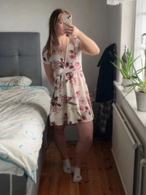Vit blommig omlottklänning - Söt kort vit klänning med rosa och bruna blommor. Omlottmodell med v-ringning och knyt i midjan. Perfekt för varma dagar och har korta ärmar. Lätt och luftig känsla.