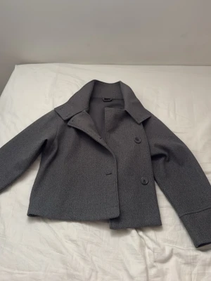Grå kort rock från Gina Tricot - Snygg grå kort rock från Gina Tricot med bred krage och dubbelknäppning framtill. Rocken har en rak passform och är tillverkad i ett mjukt, filtliknande material som ger en stilren look. Perfekt för dig som gillar minimalistisk och trendig stil.