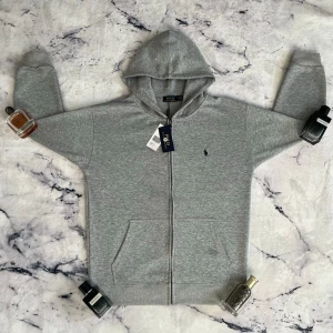 Grå zip hoodie från Polo Ralph Lauren - Klassisk grå hoodie med dragkedja från Polo Ralph Lauren. Tröjan har huva, två fickor framtill och den ikoniska lilla broderade hästloggan på bröstet. Tillverkad i mjukt bomullsmaterial som är perfekt för chill dagar eller när du vill ha en clean look.