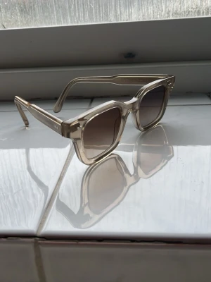  Solglasögon från CHIMI 04 Ecru - Snygga solglasögon från CHIMI i modellen ”04 Ecru”. Dem har en transparent beige båge och fyrkantig form. Glasen är tonade i en mörkare nyans vilket ger en cool kontrast. Perfekta för att lyfta din stil under soliga dagar.
