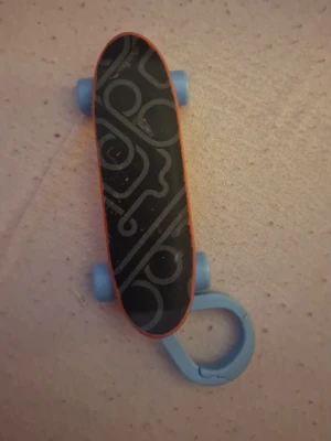 Nyckelring skateboard i plast - Cool nyckelring formad som en skateboard i plast med svart mönstrad ovansida, orange undersida och blå hjul samt blå nyckelringsfäste. Perfekt accessoar för dig som gillar skatekultur och vill ha något unikt till dina nycklar eller väska. Pris kan diskuteras