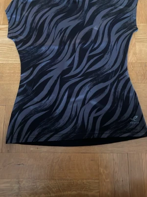 Svart zebramönstrad t-shirt  - Sportig svart t-shirt från Röhnisch med grått zebramönster på framsidan och övre delen av ryggen. Tillverkad i stretchigt syntetmaterial som andas, perfekt för träning. Kortärmad och figurnära modell med diskret logga nere till höger.