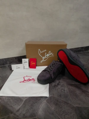 Christian Louboutin grå sneakers - Snygga svarta sneakers från Christian Louboutin med ikonisk röd sula. Skorna har en klassisk låg modell med snörning och är tillverkade i mocka. Perfekta för dig som vill sticka ut med exklusiva detaljer och lyxig känsla.