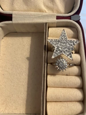Två silvriga ringar med stenar - Säljer två silvriga ringar, en med stor stjärnform täckt av gnistrande stenar och en mindre ring med rund, sol-liknande form och små stenar, som även är en snurr ring som avlägsnas stress.. Perfekta för dig som gillar statement-smycken och vill sticka ut med cool design.