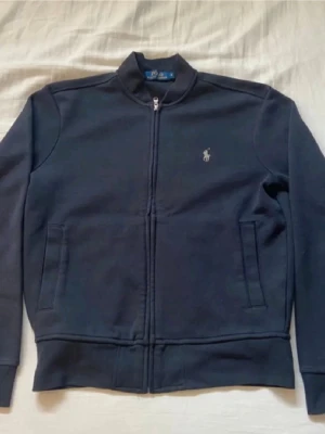 Mörkblå ziptröja från Polo Ralph Lauren - Snygg mörkblå ziptröja från Polo Ralph Lauren med klassisk ståkrage och dragkedja framtill. Tröjan har två fickor och den ikoniska broderade loggan på bröstet. Perfekt för en stilren och avslappnad look. Skick 8/10 lite nopprig men borde gå att ta bort med en nopp borttagare