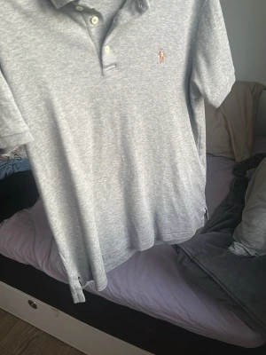Grå pikétröja från Polo Ralph Lauren - Säljer en grå pikétröja från Polo Ralph Lauren i slim fit-modell. Tröjan har klassisk krage, knappar framtill och den ikoniska broderade loggan på bröstet. Tillverkad i mjuk bomull som känns skön mot huden.