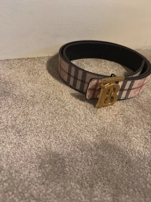 Randigt Burberry bälte med guldspänne - Snyggt bälte från Burberry med klassiskt beige, svart och rött randigt mönster. De kan även vara svart med fin färg! Perfekt accessoar för att lyfta din outfit.