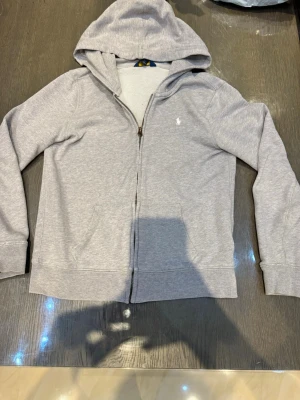 Grå zip hoodie från Ralph Lauren  - Snygg grå hoodie med dragkedja från Polo Ralph Lauren. Klassisk design med huva, fickor framtill och den ikoniska broderade loggan på bröstet. Tillverkad i mjukt bomullsmaterial som är perfekt för chill dagar. Enkel att matcha med jeans eller joggers. Litet sytt hål vid  armslutet. För 16 år XL/S. Köpt från kidsbrandstore.