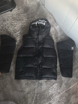Svart dunväst med huva från Burberry - Snygg svart dunväst från Burberry med avtagbara ärmar och huva. Västen har klassiskt rutigt foder i huvan och dragkedja framtill. Perfekt för lager-på-lager och streetstyle. Ikonisk logga på ena ärmen. Priset är inte hugget i sten och kan diskuteras.