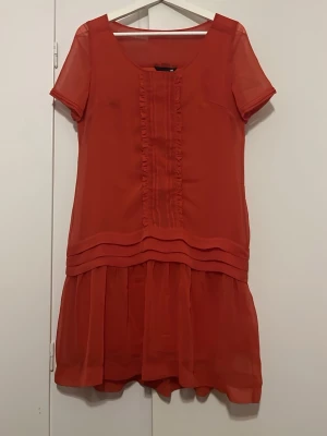 Röd kort klänning med volangdetaljer - Snygg röd kortklänning med volangdetaljer framtill och på kjolen. Klänningen har rund halsringning och korta ärmar. Tillverkad i ett lätt och luftigt material som ger en skön känsla. Perfekt för dig som vill ha luftig, fin klänning för sommaren. 