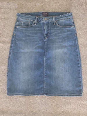 Blå jeanskjol från NYDJ - Klassisk blå jeanskjol från NYDJ med femficksmodell och knapp- samt dragkedjestängning framtill. Kjolen har en rak passform och är tillverkad i mjuk denim med lätt tvättad look. Perfekt för en avslappnad och trendig stil.