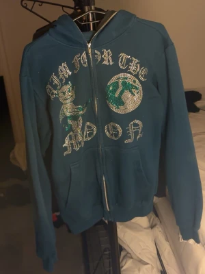 Blå hoodie med strasstryck - Blå hoodie med dragkedja och huva. Framsidan har ett stort strasstryck med texten 'Aim for the moon', en tecknad figur och en jordglob i glitter. Perfekt för dig som gillar unika och statement-plagg.