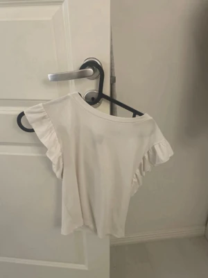 Vit topp med volangärmar - Söt vit topp med korta volangärmar och rund halsringning. Perfekt för en fräsch och enkel look. Tunn och mjuk i materialet, passar bra till jeans eller kjol.