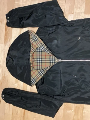 Burberry reversible rutig vindjacka - Stilren vindjacka från Burberry med klassiskt beige rutmönster och svart insida. Jackan har huva, dragkedja framtill och två fickor med dragkedja. Materialet är lätt och glansigt, perfekt för blåsiga dagar och funkar även för varma dagar. Ikoniskt Burberry-mönster på utsidan och svart på insidan den går även att vända ut och in