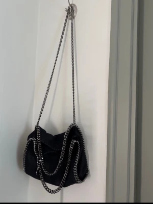 Svart axelväska Stella McCartney - Snygg svart axelväska från Stella McCartney med kedjedetaljer i silver runt kanterna och som axelrem. Väskan har en mjuk, slouchy form och är gjord i veganskt material. Perfekt för dig som vill ha en trendig och stilren väska med coola detaljer.