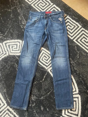 Blå raka jeans från Replay - Snygga blå jeans från Replay med raka ben och klassisk femficksmodell. Jeansen har slitningar och ljusare partier framtill för en cool, avslappnad look. Märkesdetaljer syns på fickan och insidan av linningen. Perfekta för dig som gillar streetstil.