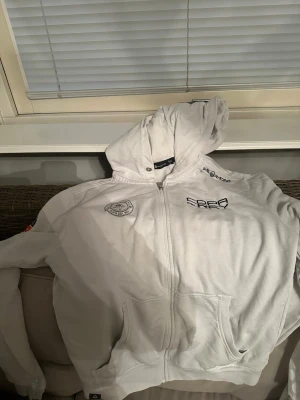Vit Sail Racing zip-hoodie - Snygg vit hoodie med dragkedja från Real Club Nautico Palma. Broderade loggor på bröstet och axeln, texten 'Copa del Rey' och 'RCNP'. Utrustad med huva och två fickor framtill. Perfekt för en sportig och avslappnad stil.