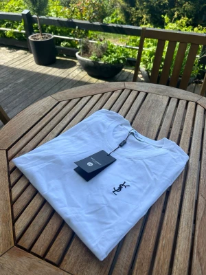 Vit t-shirt från Saint Laurent - Säljer en fräsch vit t-shirt från Saint Laurent med klassisk rund hals och svart YSL-logga broderad på bröstet. T-shirten är i mjuk bomull och har en clean, stilren look som passar perfekt till jeans eller shorts.