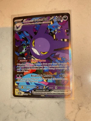 Team Rocket's Crobat EX Pokémonkort - Säljer ett holografiskt Team Rocket's Crobat EX Pokémonkort med lila och svarta färger. Kortet har coola illustrationer och detaljerad text med attacker som Biting Spree och Assassin's Return. Perfekt för samlare eller Pokémon-fans som vill utöka sin kortsamling.