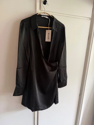 Svart satin klänning från NA-KD - Stilren svart kort klänning från NA-KD i glansigt satinmaterial. Klänningen har lång ärm, skjortkrage och en snygg omlott-drapering framtill. Perfekt för dig som gillar trendiga och eleganta looks.