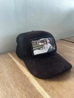 Svart maggiore keps med tryck på marlyn monroe  - Svart maggiore keps med mesh på sidorna och ett stort retroinspirerat tygmärke framtill som föreställer en kvinna i vit klänning. Kepsen har böjd skärm och justerbar baksida. Perfekt för en cool och avslappnad stil.