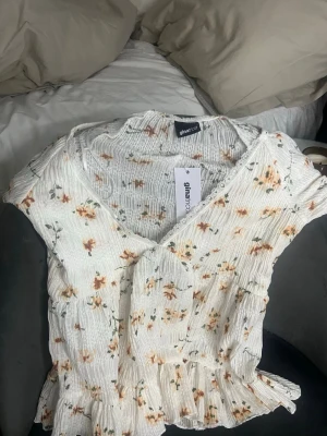 Blommig topp från Gina Tricot - Supersöt blommig topp från Gina Tricot i vit med små orange och gröna blommor. Toppen har v-ringning, korta ärmar och volangkant nedtill. Materialet är ribbad och stretchig polyester som ger en skön passform. Perfekt för vår och sommar!